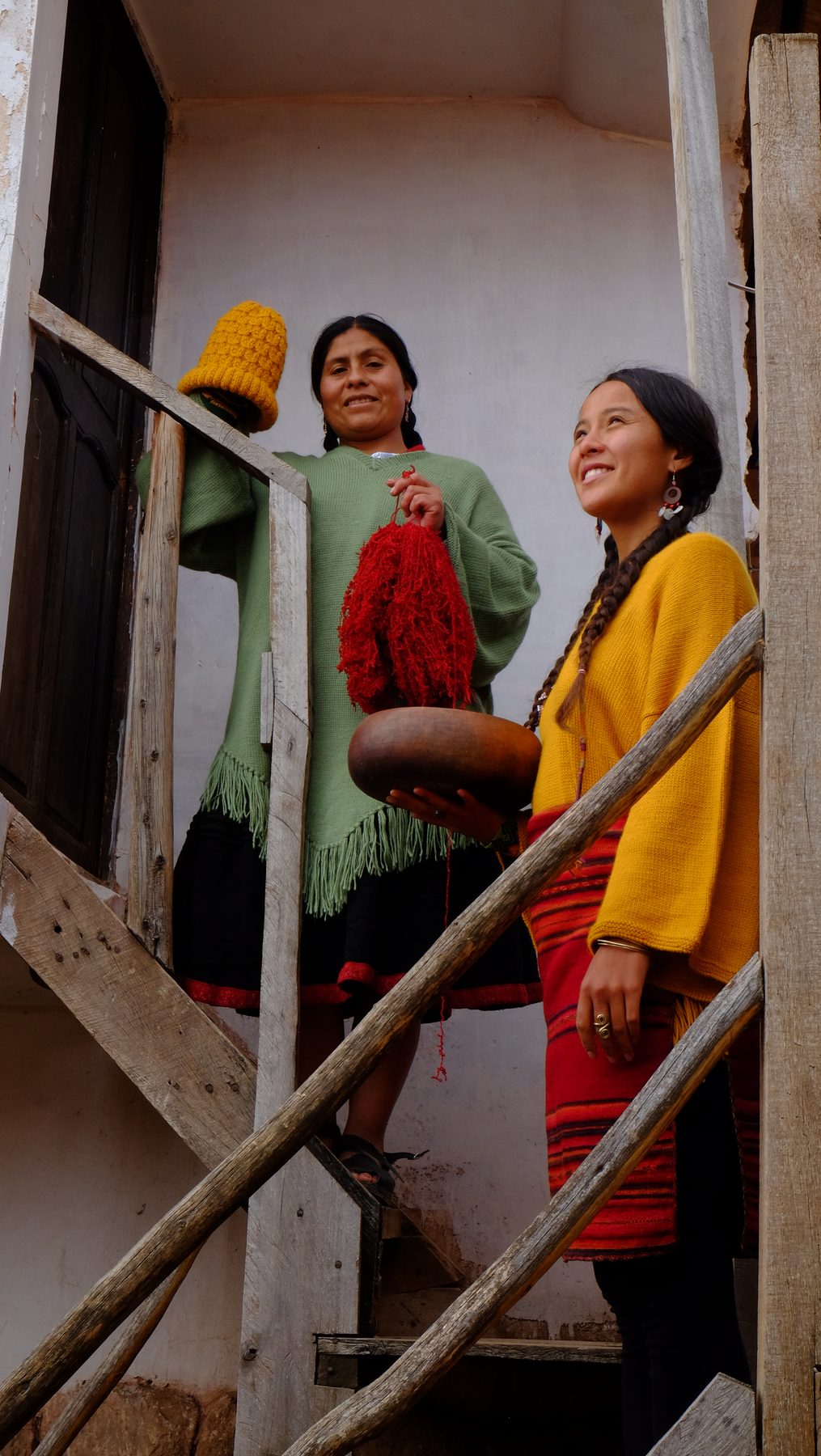 A weaver in an orange handwoven shawl, holding a spindle of red yarn against a warm earthen wall